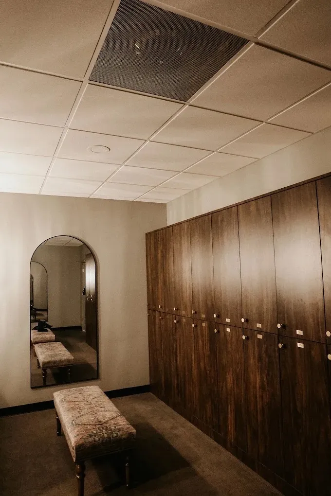 The members' locker room at Wellness Experience Pilates, with dark wood lockers, a cushioned bench, and an arched mirror.