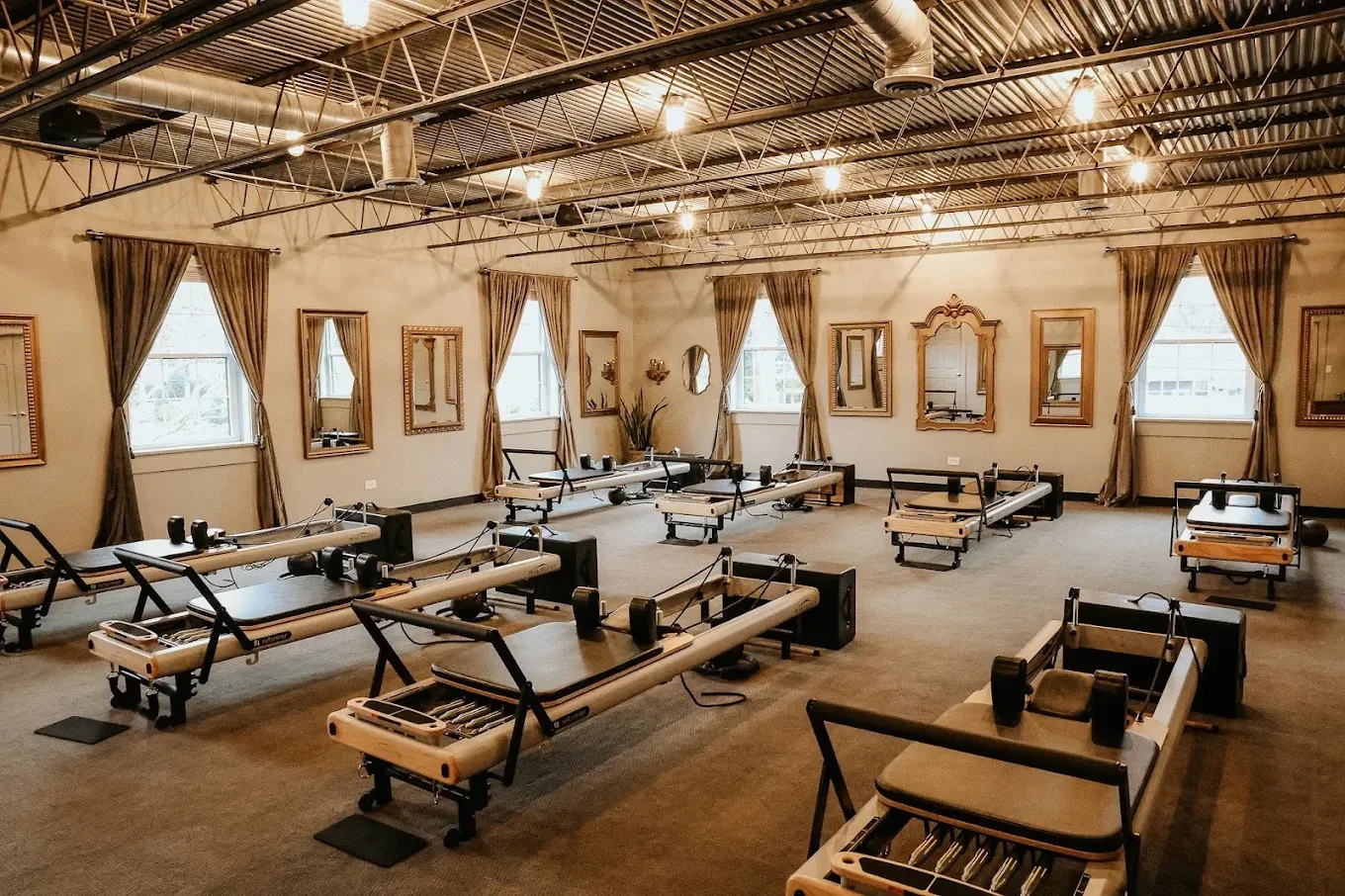 The reformer Pilates studio floor at Wellness Experience Pilates — rows of reformers under an open industrial ceiling, framed by curtained windows and gilt mirrors.