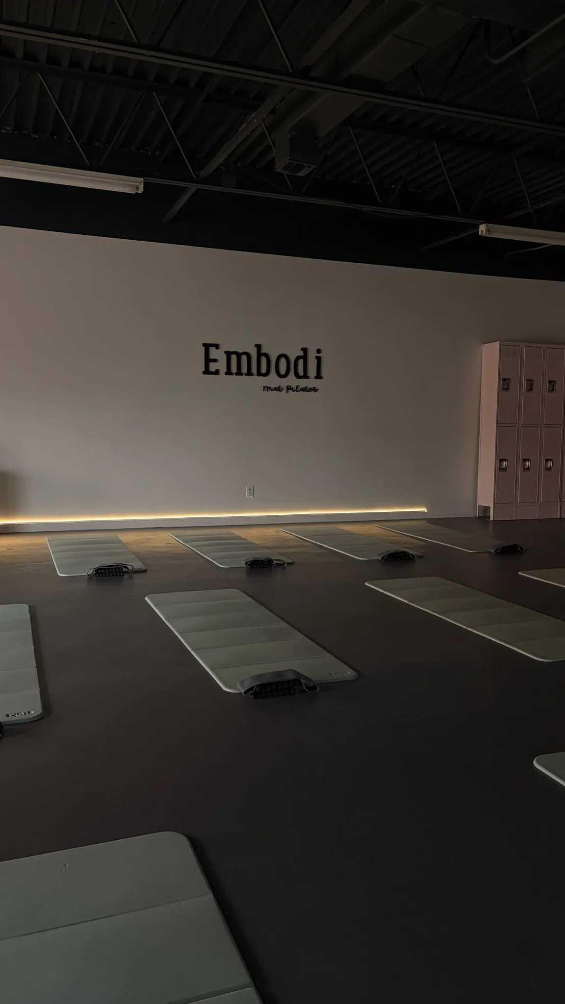 The Embodi Mat Pilates studio floor — rows of mats laid out beneath the backlit 'Embodi mat pilates' wall sign.